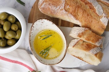 Tasty baguette served with oil, rosemary and olives on white marble table, flat lay