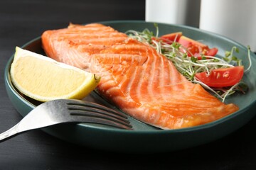 Piece of tasty grilled salmon, lemon, tomato slices and microgreens served on black wooden table, closeup
