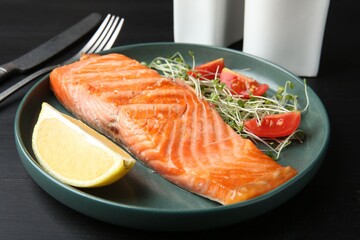 Piece of tasty grilled salmon, lemon, tomato slices and microgreens served on black wooden table, closeup