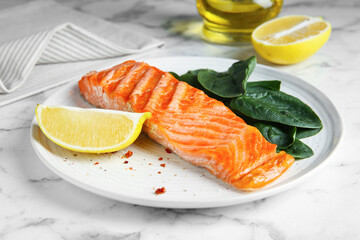 Piece of tasty grilled salmon, lemon slice and spinach served on white marble table, closeup