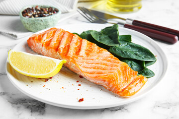 Piece of tasty grilled salmon, lemon slice and spinach served on white marble table, closeup