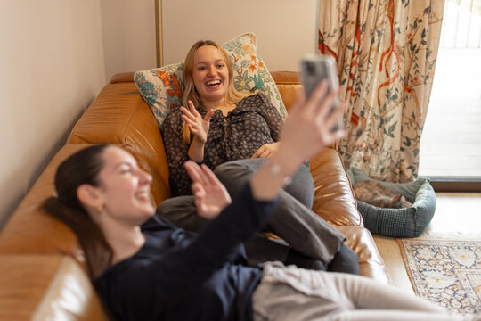 Two friends laughing and waving while taking a selfie on a leather sofa. Concept of bonding, joy, friendship and social media connection. Cat resting in pet bed in background. Horizontal, side view.