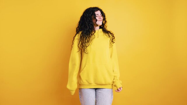 Joyful beautiful young woman with long curly hair wearing a yellow hoodie and jeans, happily dancing and spinning against a solid yellow background