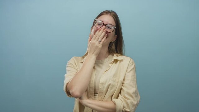 Young blonde woman wearing glasses and beige shirt covers mouth with hand and smiles in studio; joy.