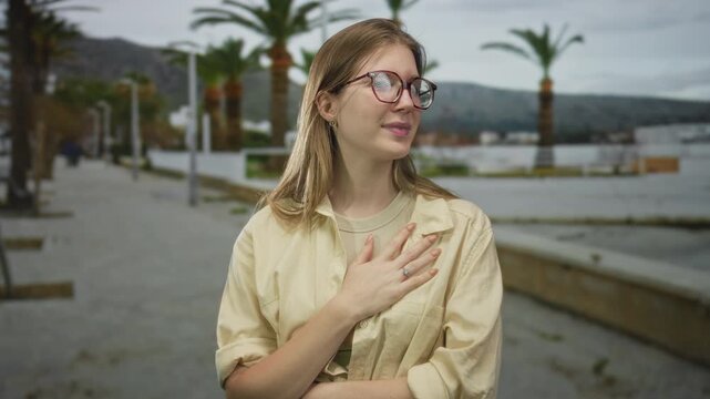 Woman with glasses and beige shirt holds hand on chest walking along palm lined street by waterfront; pain.