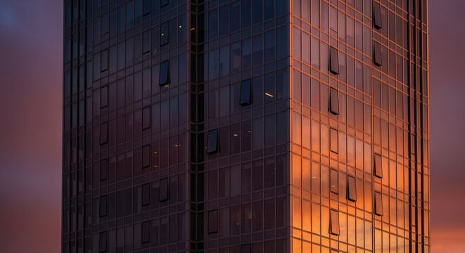 Exterior of glass building, some windows are open, reflecting orange sky, representing ambition, success, and modern business or residence