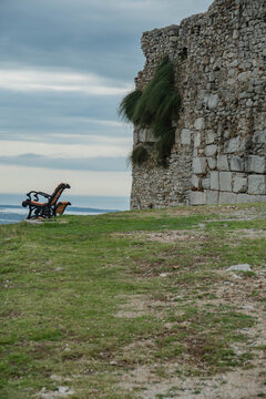 A tranquil, moody, vertical shot capturing a single, empty wooden park bench positioned on a grassy hill next to the imposing, ancient stone wall of Lezha Castle (Kalaja e Lezh&euml;s).