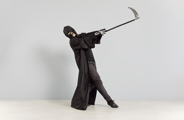 Person wearing a Death costume on Halloween. Young man in a black cloak, skull mask and bone gloves holding a scythe and dancing in a funny way by a light gray wall color background