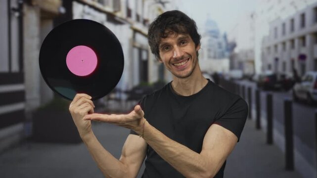 Man holding pink label vinyl record with open palm presentation gesture on city street scene, smiling at disc; nostalgia.