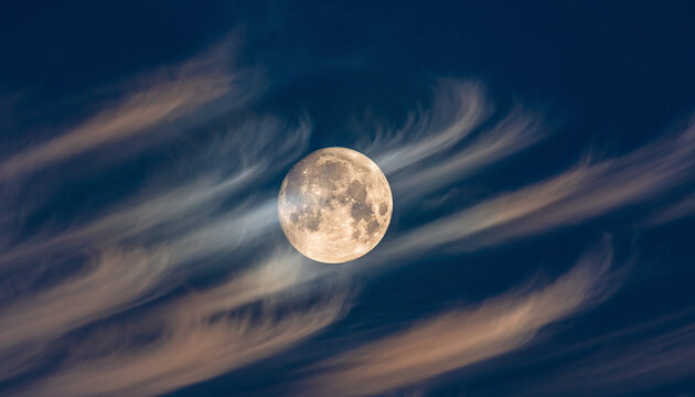 Full moon shining through wispy clouds against a dark blue sky.