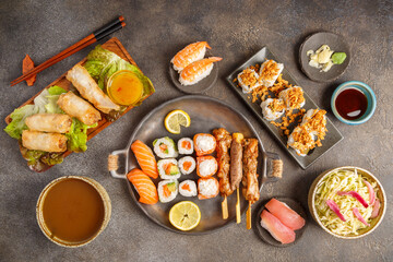 Assortiment de plats japonais : sushi, maki, California, gyozas, brochettes et salades sur table foncée avec sauces, baguettes. Vaisselle japonaise. 