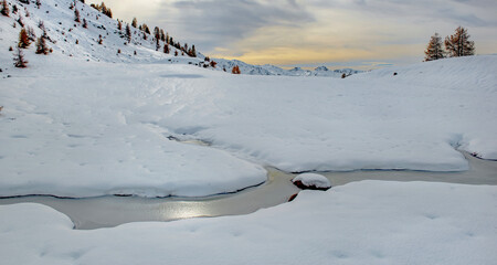 winter landscape in the mountains with hills under fresh snow and a frozen stream under a cloudy and colorful sky at sunset