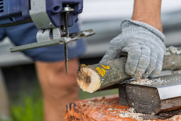 A person in work gloves uses a blue electric jigsaw to cut a tree branch. Wood shavings fly as the...