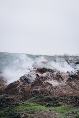 A rugged hillside emits white smoke from multiple cracks in the earth, creating a dramatic and surreal atmosphere.