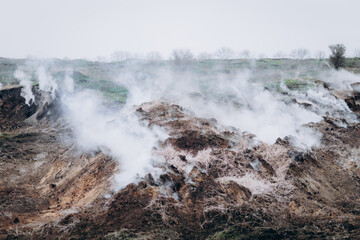 A rugged hillside emits white smoke from multiple cracks in the earth, creating a dramatic and surreal atmosphere.