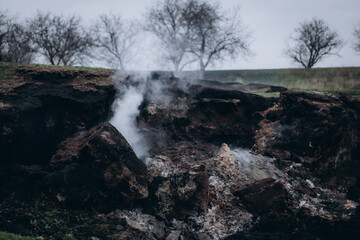 A rugged hillside emits white smoke from multiple cracks in the earth, creating a dramatic and surreal atmosphere.