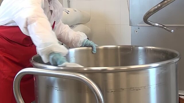 In a professional kitchen, someone wearing protective clothing cleans the inside of a large, round, stainless steel mixing bowl with a white cloth. The surface is wiped carefully.