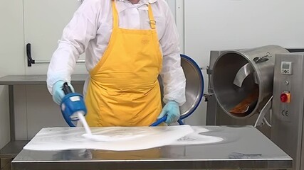 A worker in a white suit and yellow apron sanitizes a stainless steel table with a cleaning solution using a sprayer, next to a food processing machine in a kitchen.