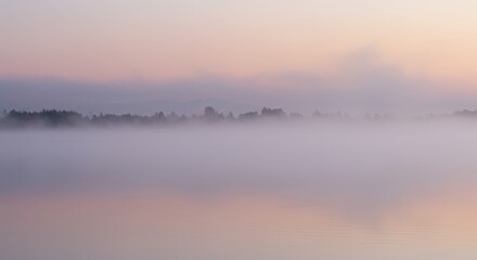 Fototapeta premium Tranquil water surface reflects soft pastel colors during a heavily misty sunrise or sunset over a distant tree line.