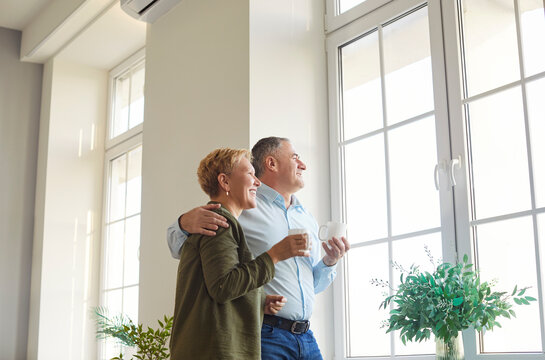 Happy senior couple, mature husband and wife standing looking at new frame window in comfortable living room to enjoy tea, talking, smiling relax at home on leisure weekend, planning future life