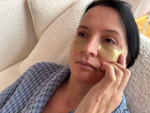 Mature woman with fine wrinkles applying under-eye patches for skin hydration. Anti-aging skincare routine, eye area care, moisturizing cosmetic treatment at home