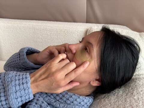 Mature woman with fine wrinkles applying under-eye patches for skin hydration. Anti-aging skincare routine, eye area care, moisturizing cosmetic treatment at home