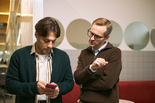 Caucasian young adult man using smartphone while standing next to Caucasian middle aged man gesturing and explaining business concept in modern office environment - Powered by Adobe