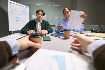 Caucasian young adult man and Black young adult woman sitting at conference table discussing...