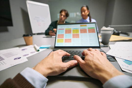 Caucasian young adult man working on laptop with project management software while multiethnic young adult colleagues discussing business strategy in modern office meeting room