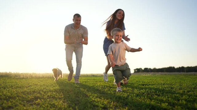 A child runs on grass followed by a man and woman, while a small dog follows too. The child leads as the man and woman stay close on the sunny field.