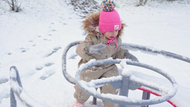 A little child girl in winter clothes rides a carousel on a playground in winter with snow