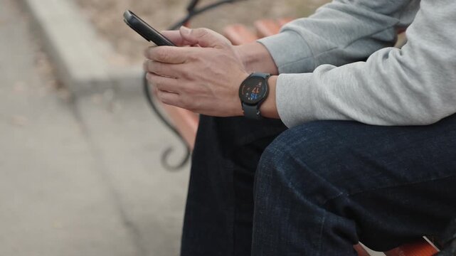 Man dressed casually in grey sweatshirt and blue jeans wearing smart watch sits on bench in peaceful park focused on his smartphone. Male person chatting online using mobile phone outdoors.