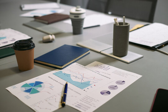 Closeup of business documents with charts and graphs lying on office table surrounded by notebooks, reusable coffee cups, pens and pencils, showing financial analysis in progress - Powered by Adobe