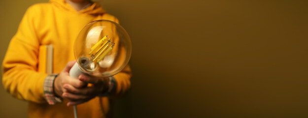 Person in yellow hoodie holding large glowing vintage-style LED light bulb against warm background. Hand holding a filament light bulb isolation on dark yellow background. Innovation concept