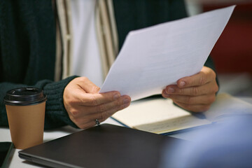 Caucasian middle aged man holding documents and reviewing paperwork at desk with disposable coffee cup and closed laptop visible, business context suggesting office environment