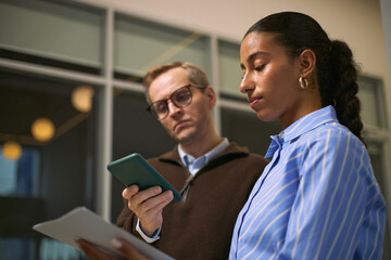 Caucasian middle aged man holding smartphone standing next to young adult Black woman reviewing documents in modern office setting collaborating on business project