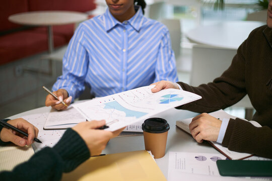 Multiethnic group of young adult professionals collaborating at table, analyzing financial charts and graphs, exchanging documents, discussing business strategy during meeting