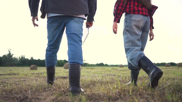 A man and woman farmers in jeans walk through a grassy field, wearing jeans and boots. The man and woman stride confidently with jeans against a backdrop of hay bales.