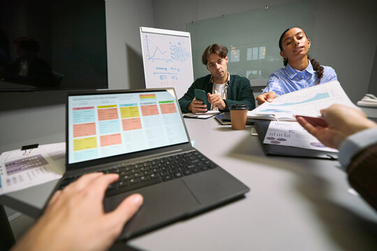 Young adult Caucasian man and young adult Black woman participating in business meeting, discussing charts and data with colleagues, laptop and documents on table, office setting - Powered by Adobe