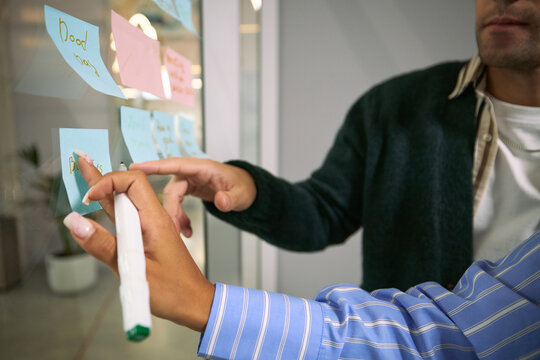 Caucasian young adult man and Black young adult woman collaborating by placing sticky notes on glass wall during business meeting, hands and partial faces visible, brainstorming ideas
