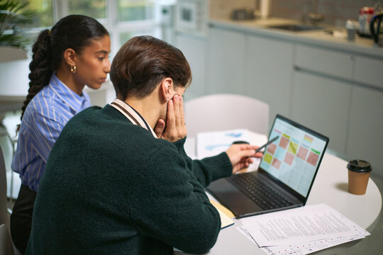 Young adult Caucasian man and young adult Black woman analyzing business data on laptop in modern office setting, discussing charts and documents, coffee cup on table