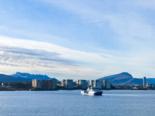 Passenger ferry approaching the modern city coastline, with snow-capped mountains and cloudy sky in the background