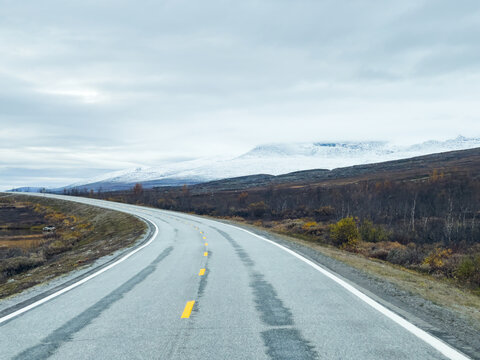 Desolate winter landscape with an asphalt road leading towards snow-capped mountains under a cloudy sky