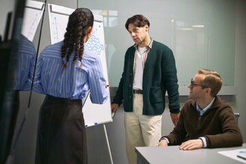 Young Caucasian woman presenting business strategy on flip chart to young Caucasian man and young Caucasian man sitting at table, colleagues discussing project in office meeting room