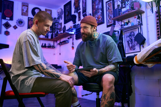 Caucasian young adult man with tattoos sitting and listening while middle aged man with tattoos and beard explaining something using digital tablet in tattoo studio
