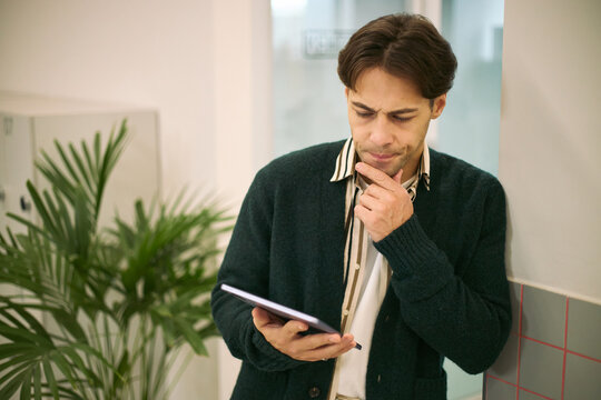 Young adult Caucasian man standing in office environment holding digital tablet, touching chin and concentrating on screen, engaging in business decision making process
