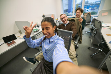 Young adult Hispanic woman taking selfie while smiling and making peace sign, two young adult Caucasian men sitting on office chairs behind her laughing and posing in modern business office
