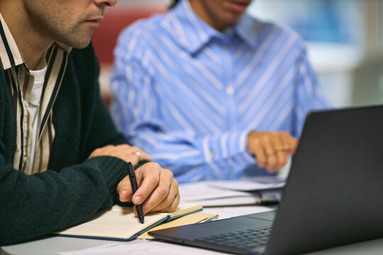 Caucasian young adult man and Black young adult woman collaborating at desk, reviewing documents and taking notes while working on laptop in modern business environment - Powered by Adobe