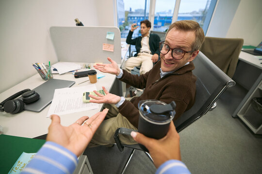 Caucasian middle aged man sitting at office desk gesturing with hands while looking at Black woman holding reusable cup, young Caucasian man sitting in background talking on phone