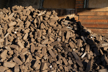 A large pile of brown bricks sits stacked in a construction area beside a wooden wall, illuminated by bright daylight